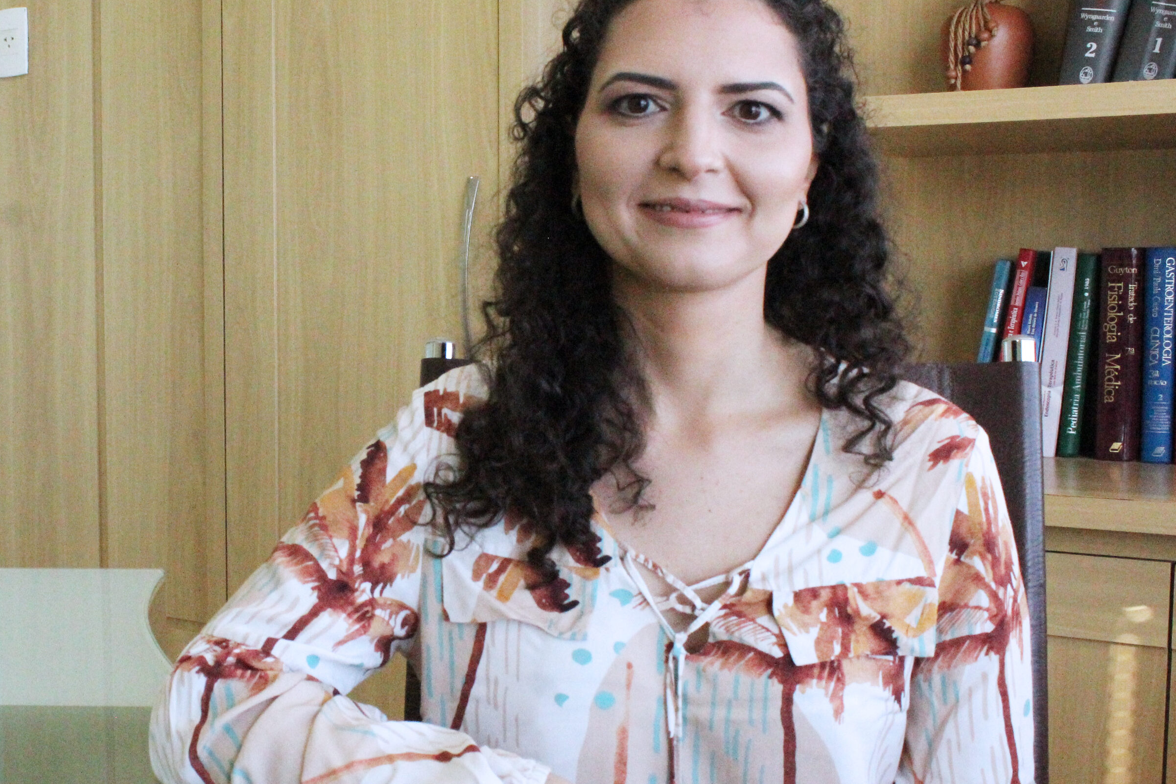 médica gastroenterologista com cabelos cacheados e escuros, vestindo uma blusa estampada em tons de marrom e azul claro, está sentada à mesa sorrindo. Ao fundo, há uma estante de madeira com livros e objetos decorativos, incluindo volumes sobre medicina e fisiologia. O ambiente é bem iluminado e transmite um clima profissional e acolhedor.